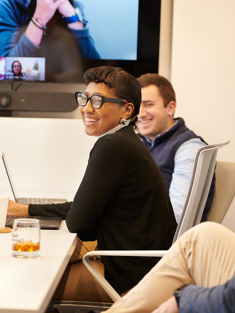 A female and a male office employee smile while sitting at conference table. Other employees join the meeting from a video conference screen.
