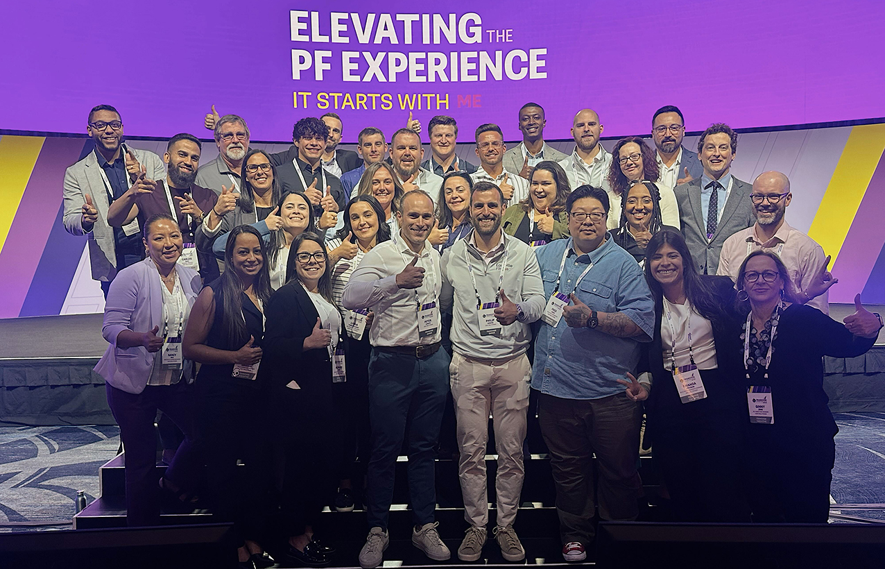 A group of Easy Mile Fitness team members gather on a stage in front of a screen that reads "Elevating the PF Experience" in large letters followed by "It starts with me" in a smaller font size.