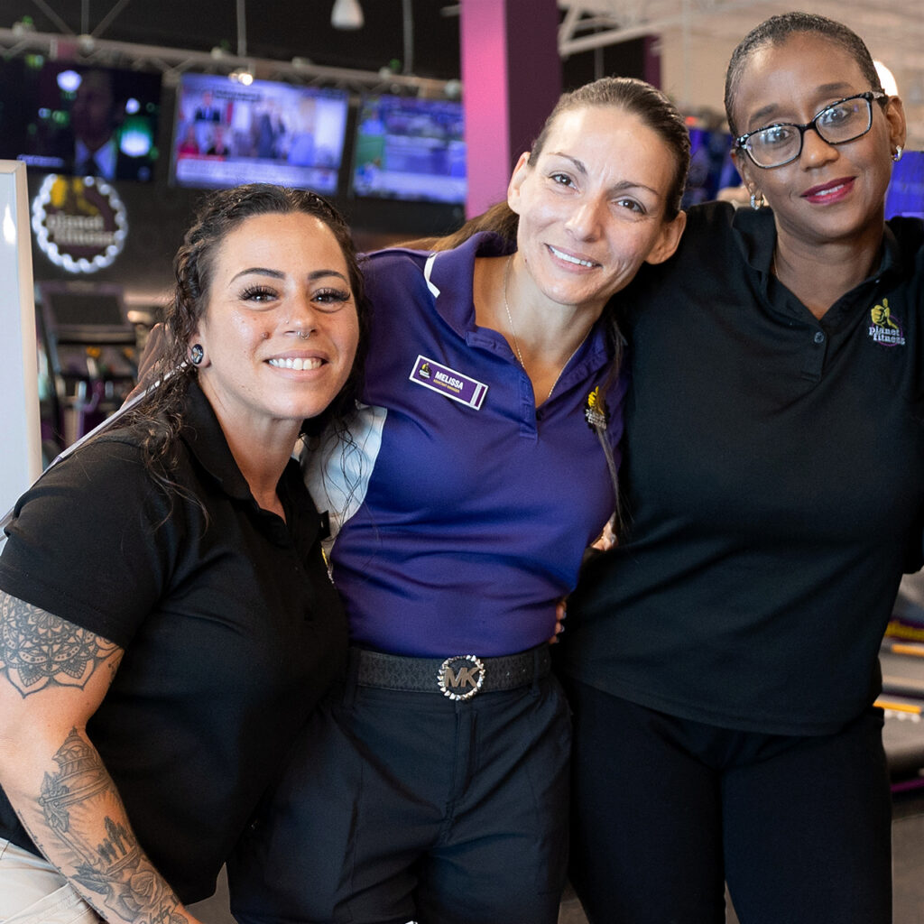 Three women wearing Planet Fitness staff shirts stand in a Planet Fitness club with their arms wrapped behind each other.