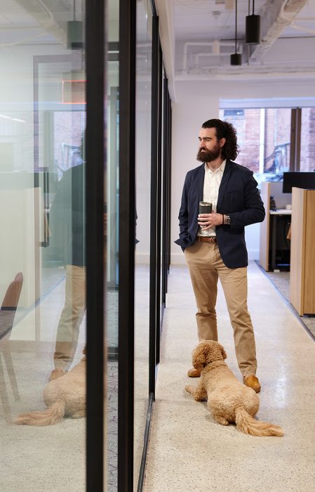 careers_hero_office-174_820x1276 An officer worker stands outside the entrance to a conference room with glass walls. He holds a thermos. A dog lies at his feet.