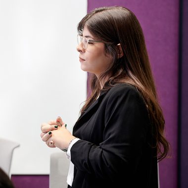 our-values_hero_office-1_600x600 A female business woman clasps her hands during a presentation. She stands next to a white board and purple wall.