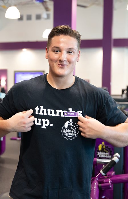 our-values_hero_pf_820x1276 An EMF employee stands in a Planet Fitness club. He points to his tshirt that reads "thumbs up."